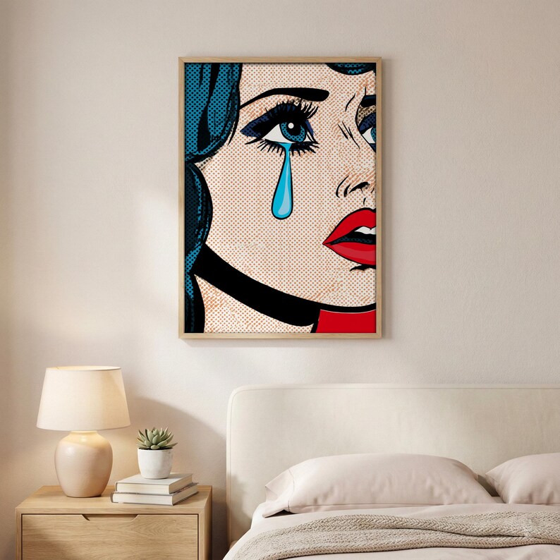 May include: Pop art print of a crying woman with blue hair and a tear. The artwork features bold lines, a red lip, and a black choker. The print is framed in a light wood frame and hangs on a white wall.