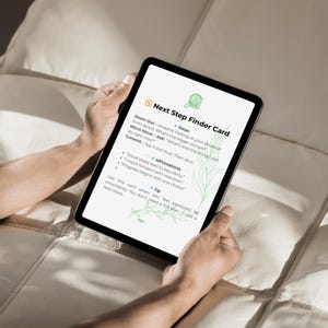May include: A black tablet showing a "Next Step Finder Card" with text including "Zoom Out," "Micro Move," and "Commit." The card also lists affirmations and a tip for overcoming uncertainty. The tablet is held by hands.