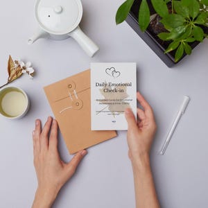 Puede incluir: Una persona sostiene una tarjeta blanca con el texto "Daily Emotional Check-in". Tambi&eacute;n se ven un sobre marr&oacute;n, una tetera blanca, una taza de t&eacute;, un bol&iacute;grafo y una planta en una maceta negra.