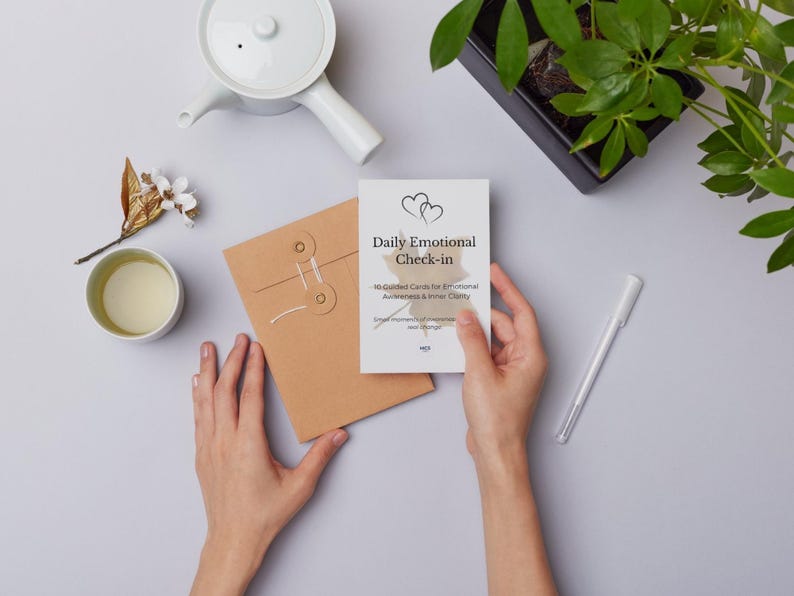 Puede incluir: Una persona sostiene una tarjeta blanca con el texto "Daily Emotional Check-in". Tambi&eacute;n se ven un sobre marr&oacute;n, una tetera blanca, una taza de t&eacute;, un bol&iacute;grafo y una planta en una maceta negra.