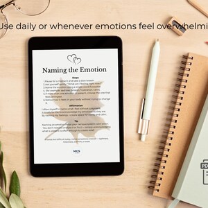 May include: A digital tablet shows a document titled "Naming the Emotion" with steps and affirmations. A pen, notebooks, and glasses are on a light wood surface. The text "Use daily or whenever emotions feel overwhelming" is at the top.