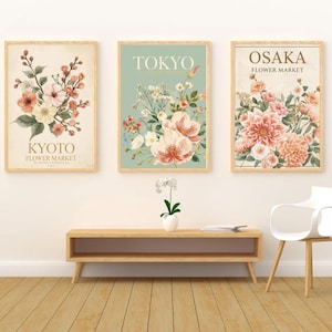 以下が含まれることがあります： 3つの額入りフラワーアートプリント。プリントには、京都フラワーマーケット、東京、大阪フラワーマーケットと書かれた花の水彩イラストが描かれています。フレームは薄茶色で、背景は薄いベージュ色です。
