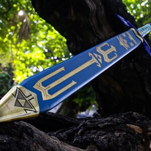 May include: A blue and gold decorative sword with a trident design. The sword features a gold blade tip and a blue handle. The sword is resting on a dark, textured surface, with a blurred green background.