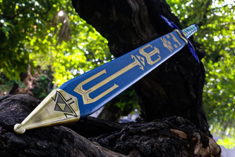 May include: A blue and gold decorative sword with a trident design. The sword features a gold blade tip and a blue handle. The sword is resting on a dark, textured surface, with a blurred green background.