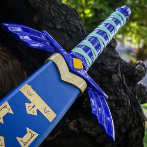 May include: A blue and gold fantasy sword prop, featuring a detailed design with a blue blade and hilt. The hilt is wrapped in light blue and green bands. The sword rests against a textured, dark brown surface.