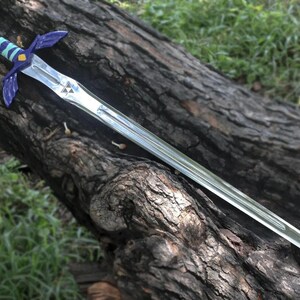 May include: A silver-colored fantasy sword with a blue and green handle and guard, resting on a weathered log. The sword features a triangular emblem on the blade and a pointed tip. The handle is wrapped in a striped pattern.