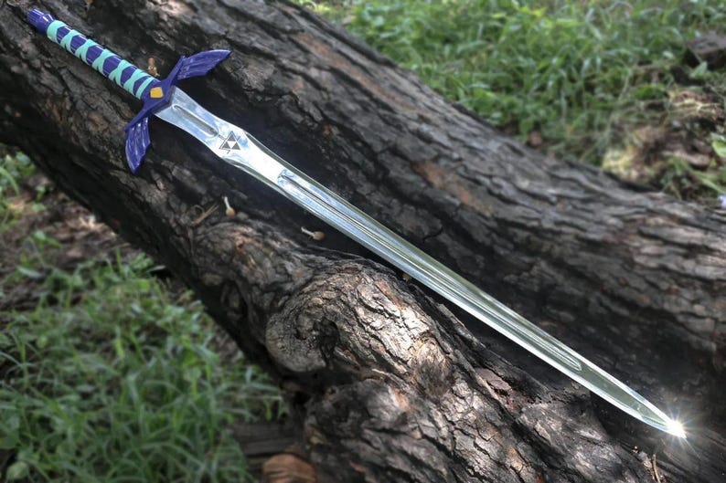 May include: A silver-colored fantasy sword with a blue and green handle and guard, resting on a weathered log. The sword features a triangular emblem on the blade and a pointed tip. The handle is wrapped in a striped pattern.