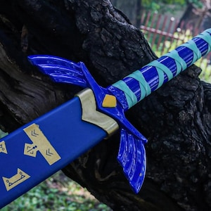 May include: A blue and gold fantasy sword with a wrapped handle. The sword features a blue blade with gold accents and a crossguard with wing-like designs. The handle is wrapped in blue and teal tape. The sword is resting on a tree trunk.