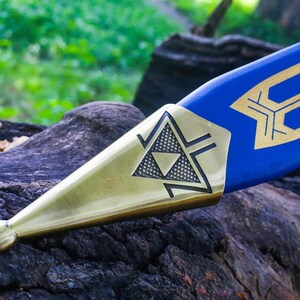 May include: Close-up of a blue and gold sword. The sword features a blue blade with gold geometric designs. The hilt is gold with a black triangle design. The sword is resting on a dark, textured surface.