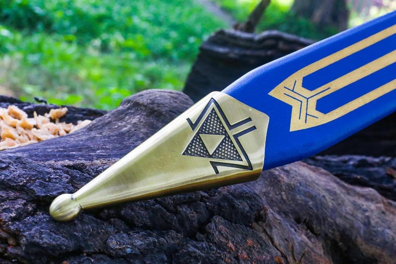 May include: Close-up of a blue and gold sword. The sword features a blue blade with gold geometric designs. The hilt is gold with a black triangle design. The sword is resting on a dark, textured surface.