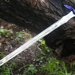 May include: A silver-colored sword with a blue and white handle and blue crossguard. The sword is lying on a log with green grass and foliage in the background. The sword has a long, pointed blade.
