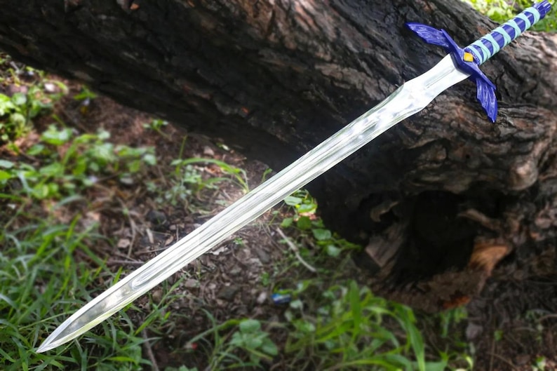 May include: A silver-colored sword with a blue and white handle and blue crossguard. The sword is lying on a log with green grass and foliage in the background. The sword has a long, pointed blade.