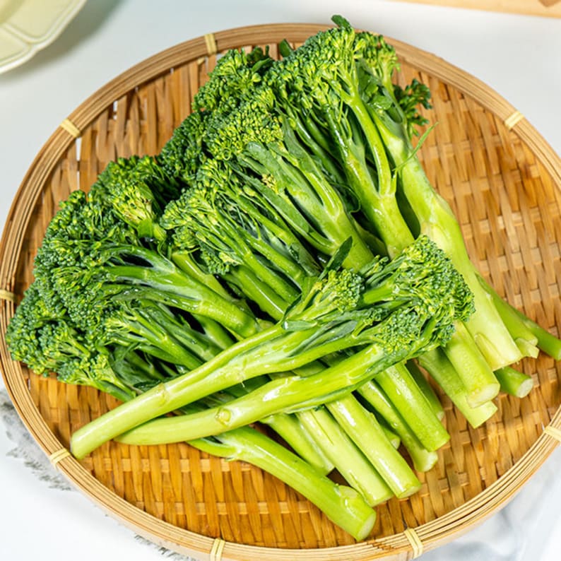 Pu&ograve; includere: Un mazzo di broccolini freschi con cimette verde brillante e gambi lunghi e sottili, disposti su un vassoio rotondo in bamb&ugrave; intrecciato. La verdura presenta boccioli compatti e steli lisci e sodi, con un aspetto naturale, crudo e croccante.