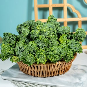 Pu&ograve; includere: Una grande e densa testa di cimette di broccoli verdi freschi disposta in un cesto rotondo di vimini intrecciato color marrone chiaro. Il broccolo presenta una superficie ruvida e irregolare di colore verde brillante, che riempie il cesto.
