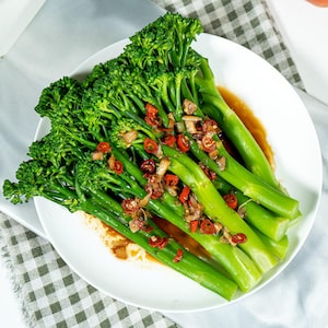 Pu&ograve; includere: Un piatto bianco riempito con gambi di broccolini verde brillante, conditi con una salsa saporita, piccoli pezzi di aglio saltato e sottili fette di peperoncino rosso. Le verdure sono disposte a grappolo, mostrando i loro lunghi gambi.