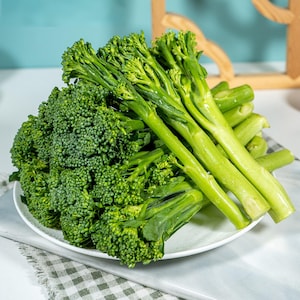 Pu&ograve; includere: Un mucchio di broccolini freschi su un piatto bianco. La verdura presenta lunghi steli sottili di colore verde chiaro, sormontati da grappoli di piccole infiorescenze testurizzate di colore verde scuro. I gambi sono lisci, sodi e sovrapposti.
