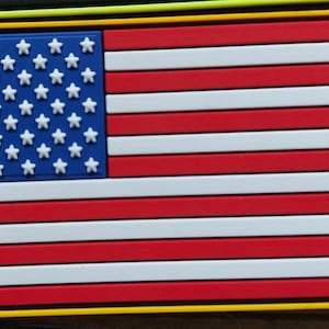 Könnte beinhalten: Rechteckiger Aufnäher mit der amerikanischen Flagge. Die Flagge hat ein blaues Feld mit weißen Sternen in der oberen linken Ecke und rot-weißen Streifen. Der Aufnäher ist mit einem gelben Rand umrandet.
