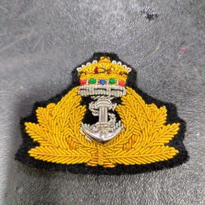 May include: A navy insignia featuring a gold crown, anchor, and laurel leaves. The emblem is embroidered with gold and silver threads, set against a black background. The crown has red, green, and blue accents.