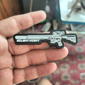 Op de afbeelding: Een zwart-wit patch in de vorm van een geweer. De patch heeft de woorden "SILENT NIGHT" in witte tekst. Het geweer heeft een vizier en andere details.