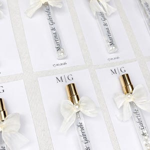 Puede incluir: Varios tubos de vidrio transparente con tapas doradas, llenos de pequeñas perlas blancas, están dispuestos sobre tarjetas blancas. Cada tubo está adornado con un lazo blanco y una perla. El texto "Martina & Gabriele" y la fecha "17.10.2026" están impresos.