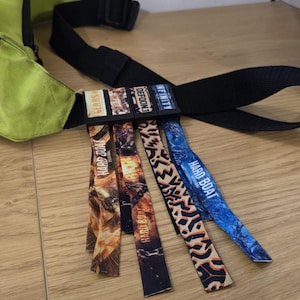 Op de afbeelding: Een limoengroene heuptas met een zwarte riem en vier bevestigde stoffen polsbandjes. De polsbandjes tonen diverse grafische ontwerpen en teksten, waaronder Gears, Defqon.1, Infinity en Hard Boat Festival in verschillende kleuren.