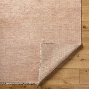 May include: A rectangular, blush-colored rug with a textured surface. A corner of the rug is folded back, revealing the underside with a lighter, striped pattern. The rug is placed on a wooden floor.