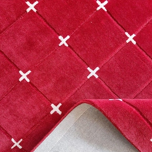 May include: A red rug with a woven design featuring white cross patterns. The rug's texture is visible, and a portion is folded back, revealing a gray backing. This decorative rug adds a pop of color and pattern to any room.