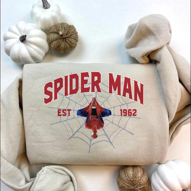 Puede incluir: Sudadera beige con las palabras "SPIDER MAN" en texto rojo arqueado sobre un gr&aacute;fico de Spider-Man colgado de una telara&ntilde;a. El texto "EST 1962" est&aacute; debajo. La sudadera est&aacute; rodeada de calabazas blancas y una bola decorativa.