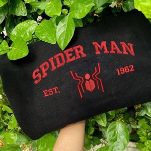 Pode incluir: Camisola preta com as palavras "SPIDER MAN" em vermelho, arqueadas na parte superior. Abaixo do texto, um gr&aacute;fico de aranha vermelha e o ano "1962". A palavra "EST." est&aacute; abaixo do texto. A camisola &eacute; rodeada por folhas verdes.