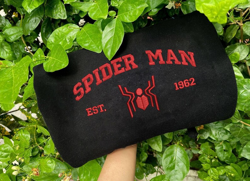 Pode incluir: Camisola preta com as palavras "SPIDER MAN" em vermelho, arqueadas na parte superior. Abaixo do texto, um gr&aacute;fico de aranha vermelha e o ano "1962". A palavra "EST." est&aacute; abaixo do texto. A camisola &eacute; rodeada por folhas verdes.