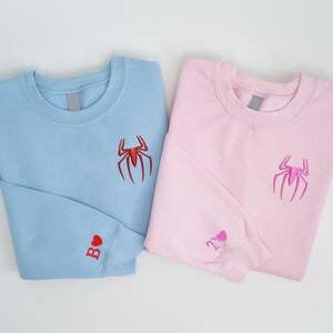 K&ouml;nnte beinhalten: Zwei Sweatshirts, eines hellblau und eines rosa, werden gezeigt. Jedes hat ein auf der Brust gesticktes Spinnenemblem. Das hellblaue Sweatshirt hat ein rotes Herz und den Buchstaben "B" auf dem &Auml;rmel. Das rosa Sweatshirt hat ein rosa Herz und den Buchstaben "T" auf dem &Auml;rmel.