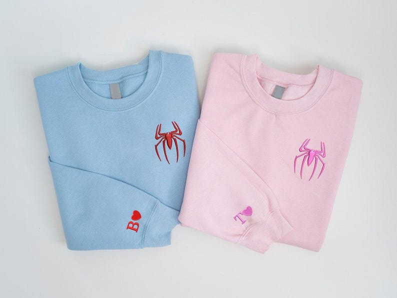 K&ouml;nnte beinhalten: Zwei Sweatshirts, eines hellblau und eines rosa, werden gezeigt. Jedes hat ein auf der Brust gesticktes Spinnenemblem. Das hellblaue Sweatshirt hat ein rotes Herz und den Buchstaben "B" auf dem &Auml;rmel. Das rosa Sweatshirt hat ein rosa Herz und den Buchstaben "T" auf dem &Auml;rmel.