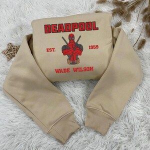 Puede incluir: Sudadera beige con la palabra "DEADPOOL" en letras rojas. La sudadera tiene un gr&aacute;fico de Deadpool con espadas cruzadas, el texto "EST. 1959" y "WADE WILSON" en rojo.