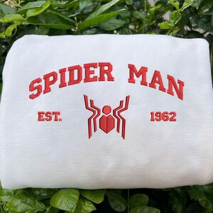 Pode incluir: Camisola branca com a inscri&ccedil;&atilde;o vermelha "SPIDER MAN" em arco. Abaixo, um emblema de aranha vermelho e o texto "EST. 1962". O fundo &eacute; composto por folhas verdes.