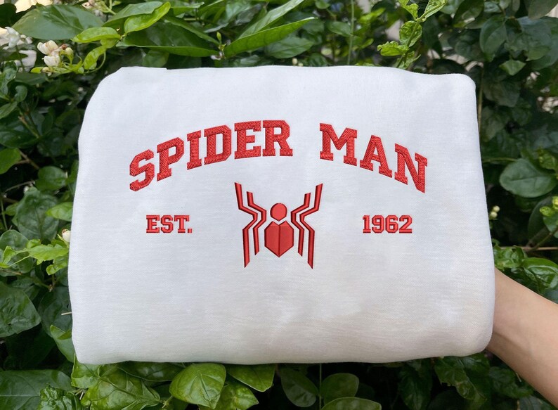 Pode incluir: Camisola branca com a inscri&ccedil;&atilde;o vermelha "SPIDER MAN" em arco. Abaixo, um emblema de aranha vermelho e o texto "EST. 1962". O fundo &eacute; composto por folhas verdes.