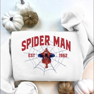 Puede incluir: Sudadera blanca con la inscripci&oacute;n roja "SPIDER MAN" sobre un dise&ntilde;o de telara&ntilde;a. El texto "EST 1962" est&aacute; debajo de la telara&ntilde;a. Una figura de Spider-Man cuelga boca abajo en el centro de la telara&ntilde;a.