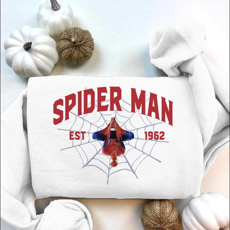 Puede incluir: Sudadera blanca con la inscripci&oacute;n roja "SPIDER MAN" sobre un dise&ntilde;o de telara&ntilde;a. El texto "EST 1962" est&aacute; debajo de la telara&ntilde;a. Una figura de Spider-Man cuelga boca abajo en el centro de la telara&ntilde;a.