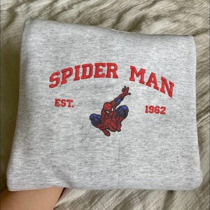 Puede incluir: Una sudadera gris claro doblada con las palabras "SPIDER MAN" en rojo, arqueadas en la parte superior, con "EST." y "1962" debajo. Un gr&aacute;fico de Spider-Man rojo y azul est&aacute; centrado debajo del texto.
