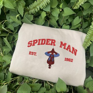 Pode incluir: Camisola bege com as palavras "SPIDER MAN" em vermelho, arqueadas sobre uma imagem do Homem-Aranha pendurado de cabe&ccedil;a para baixo. Abaixo, "EST. 1962" em vermelho. A camisola &eacute; rodeada por folhagem verde.