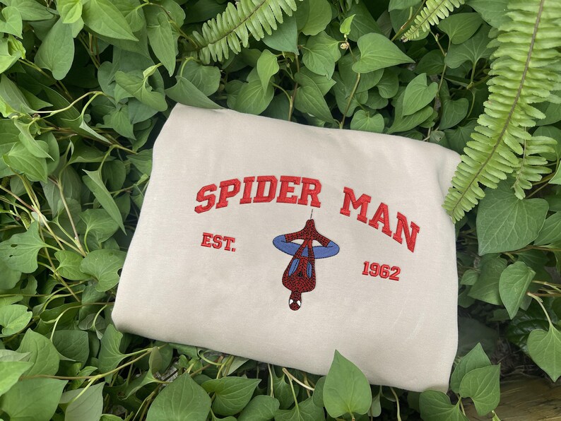 Pode incluir: Camisola bege com as palavras "SPIDER MAN" em vermelho, arqueadas sobre uma imagem do Homem-Aranha pendurado de cabe&ccedil;a para baixo. Abaixo, "EST. 1962" em vermelho. A camisola &eacute; rodeada por folhagem verde.