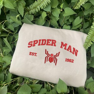 Pode incluir: Um moletom dobrado, branco sujo, com as palavras "SPIDER MAN" bordadas em vermelho, juntamente com um emblema de aranha e o ano "1962". O moletom est&aacute; rodeado por folhagem verde.