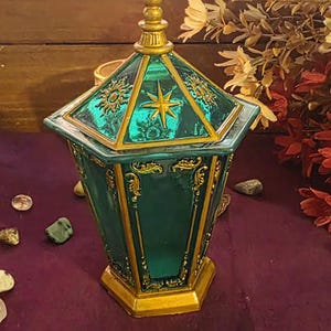 May include: A hexagonal teal glass jar with a removable lid, featuring gold-painted ornate filigree accents and a central star motif on the lid. The container has a pointed finial top and a solid gold-colored base, with a translucent teal body.