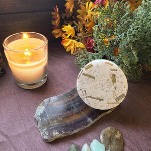 May include: A lit candle in a clear glass sits next to a white, circular item with green and gold accents, resting on a dark, multi-colored stone. Several small, colorful stones are in front. A bouquet of flowers is in the background.