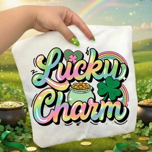 May include: A white t-shirt with the words "Lucky Charm" in a colorful, bubble-style font. The design includes a rainbow, a pot of gold, and a green shamrock, all set against a green field with a rainbow in the background.