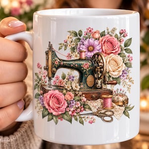 May include: A white ceramic mug featuring a graphic illustration of a vintage dark green sewing machine surrounded by a vibrant floral arrangement of pink and cream roses, purple daisies, and green leaves, with a spool of pink thread and scissors.