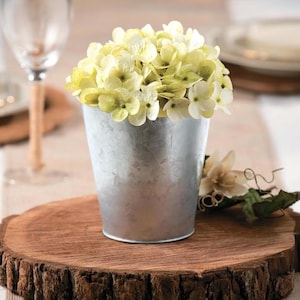 May include: A galvanized metal bucket filled with light green and white flowers sits on a rustic wooden slab. The bucket has a tapered shape and a shiny, metallic finish. The flowers are clustered together, creating a full, textured appearance.