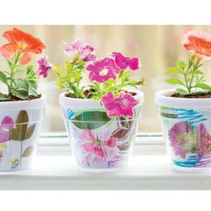 May include: Three small, decorative flower pots with colorful petunias. The pots are white with floral and abstract designs. The petunias have vibrant colors, including pink, orange, and purple. The pots are sitting on a white windowsill.