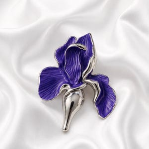 Puede incluir: Un broche con forma de flor de iris, que presenta pétalos de esmalte púrpura intenso con detalles texturizados y una base de metal plateado pulido. El diseño incluye un tallo plateado liso y redondeado con pétalos curvos en abanico.