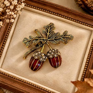 May include: A brooch featuring two acorns with deep red enamel bodies and caps encrusted with clear rhinestones, attached to a textured, antique gold-toned metal oak leaf branch with detailed veining and a stem extending from the top.