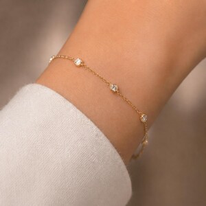 May include: A delicate gold bracelet featuring several small, round, clear gemstones. The bracelet is thin and elegant, with the gemstones evenly spaced along the chain. The jewelry is worn on a wrist, with a white sleeve in the background.